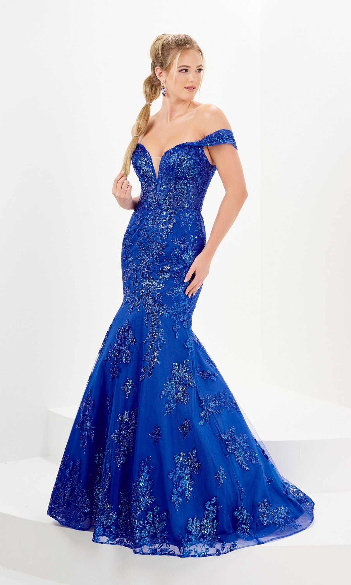 A woman stands on a white platform wearing the Tiffany Sheer Corset Glitter Mermaid Prom Dress 16080, featuring a fitted off-the-shoulder royal blue design with a sheer corset bodice and sparkling floral sequin details.
